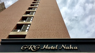 A tall building with a brick facade extending upwards against a cloudy sky. The lower part of the structure has black signage with elegant white lettering displaying 'GRG Hotel Naha'. Several rows of windows punctuate the brick exterior, creating a pattern.
