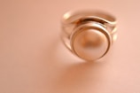 A close-up of a silver ring featuring a large, smooth pearl set in the center. The background is softly blurred in a warm, beige tone, emphasizing the elegance of the jewelry.