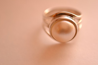 A close-up of a silver ring featuring a large, smooth pearl set in the center. The background is softly blurred in a warm, beige tone, emphasizing the elegance of the jewelry.