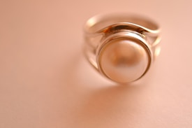 A close-up of a silver ring featuring a large, smooth pearl set in the center. The background is softly blurred in a warm, beige tone, emphasizing the elegance of the jewelry.