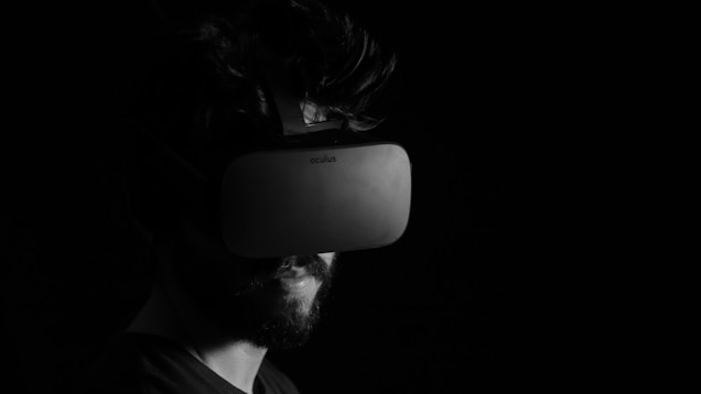 A person is wearing a virtual reality headset in a dimly lit environment. The image is in black and white, emphasizing the contrast between the dark background and the gadget. The person has a beard and short hair, and the headset partially obscures their face.