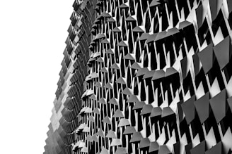 A building facade featuring a complex and geometric design with repetitive, triangular metal elements creating an intricate pattern. The play of light and shadow enhances the three-dimensional effect and adds depth.