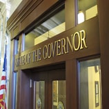 The image depicts the entrance to an office with large wooden doors and a prominent sign that reads 'Office of the Governor' in gold letters. The office is flanked by an American flag and features classical architectural elements, including columns and ornamental details. The atmosphere suggests a formal and official setting.