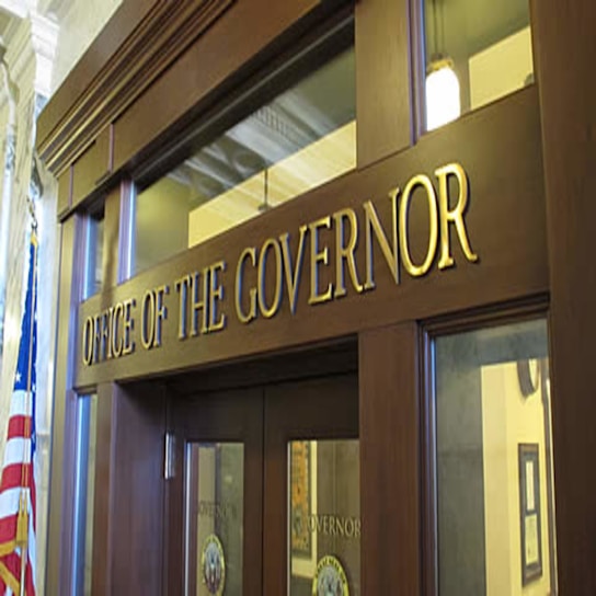 The image depicts the entrance to an office with large wooden doors and a prominent sign that reads 'Office of the Governor' in gold letters. The office is flanked by an American flag and features classical architectural elements, including columns and ornamental details. The atmosphere suggests a formal and official setting.