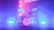 A drum set is illuminated by vibrant colored lights, with shades of pink, purple, and blue creating a dramatic stage effect. The drums, including cymbals, toms, and a bass drum with a visible logo, are positioned centrally, surrounded by a misty, smoky atmosphere that enhances the concert-like setting.