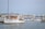 A marina filled with numerous sailboats and yachts docked in the harbor. A prominent white yacht with wooden accents is in the foreground, with the name 'Fifer' visible on its side. Several buildings line the waterfront in the background, with hills visible under a clear sky.