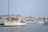 A marina filled with numerous sailboats and yachts docked in the harbor. A prominent white yacht with wooden accents is in the foreground, with the name 'Fifer' visible on its side. Several buildings line the waterfront in the background, with hills visible under a clear sky.
