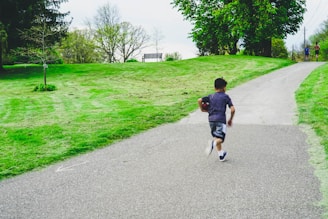 A young boy runs up a paved pathway in a park, holding a football. The area is lush with green grass and trees, and there is a bench visible on the grassy hill. In the distance, two people are walking together along the path.