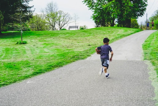 A young boy runs up a paved pathway in a park, holding a football. The area is lush with green grass and trees, and there is a bench visible on the grassy hill. In the distance, two people are walking together along the path.