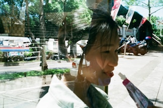 A reflection captures both an outdoor scene and a woman indoors. The outdoor scene includes greenery, a street, and parked motorbikes. The woman is seen through the reflection, seemingly holding a small tube or stick near her face.