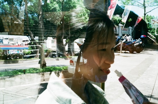 A reflection captures both an outdoor scene and a woman indoors. The outdoor scene includes greenery, a street, and parked motorbikes. The woman is seen through the reflection, seemingly holding a small tube or stick near her face.