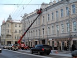A street scene with a large crane extending from a red truck, lifting a worker towards the upper facade of a historic multi-story building. The building features ornate architectural details and is lined with various small businesses at street level. Several vehicles, including cars and an SUV, are on the road, and a few pedestrians are visible on the sidewalk. The weather appears cold, with some snow accumulated on the roofs.