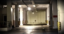 A dimly lit underground parking garage with concrete pillars, yellow and black striped bumpers, and a single overhead light casting shadows. The space seems empty and quiet, with visible reserved parking signs and some protective structures along the walls.
