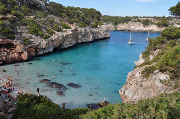 A picturesque cove featuring crystal clear turquoise water surrounded by rocky cliffs. People are swimming and relaxing on the small sandy beach, while two sailboats are anchored in the calm bay. Lush green vegetation covers the cliffs, creating a serene and inviting atmosphere.