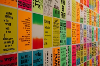 A wall covered with colorful posters featuring text in various fonts and sizes. The text appears to be a mix of poetry, event announcements, and various messages in vibrant colors. The posters are evenly arranged in a grid pattern, creating an eye-catching visual display.