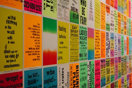 A wall covered with colorful posters featuring text in various fonts and sizes. The text appears to be a mix of poetry, event announcements, and various messages in vibrant colors. The posters are evenly arranged in a grid pattern, creating an eye-catching visual display.