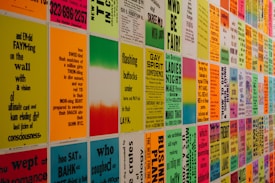 A wall covered with colorful posters featuring text in various fonts and sizes. The text appears to be a mix of poetry, event announcements, and various messages in vibrant colors. The posters are evenly arranged in a grid pattern, creating an eye-catching visual display.