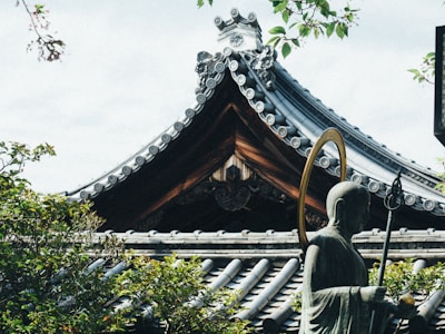 A traditional Japanese temple roof with intricate tile details is partially obscured by lush green leaves. A statue of a Buddhist monk holding a staff and a ring stands in the foreground, blending harmoniously with the natural surroundings.