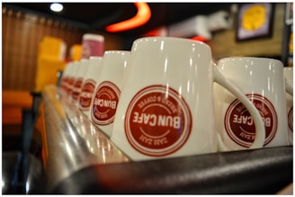 A line of white ceramic mugs with red logos arranged upside down on a metallic surface. The background is softly blurred with warm, ambient lighting, featuring some colorful, out-of-focus shapes.