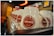 A line of white ceramic mugs with red logos arranged upside down on a metallic surface. The background is softly blurred with warm, ambient lighting, featuring some colorful, out-of-focus shapes.