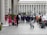 A group of bridesmaids in vibrant pink dresses and a bride in a white gown stand outside a large building with columns, while a group of groomsmen in blue suits walk towards them. Photographers are capturing the moment as passersby watch.