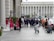 A group of bridesmaids in vibrant pink dresses and a bride in a white gown stand outside a large building with columns, while a group of groomsmen in blue suits walk towards them. Photographers are capturing the moment as passersby watch.