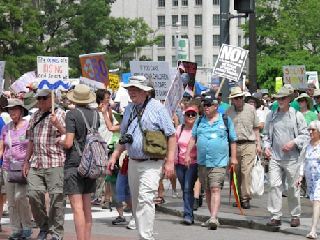 A diverse group of people are participating in a march or protest. They are holding various signs with messages related to environmental and social issues. The scene is outdoors, likely in an urban area, with trees and buildings in the background. Many participants wear sunhats or caps, suggesting a sunny day.