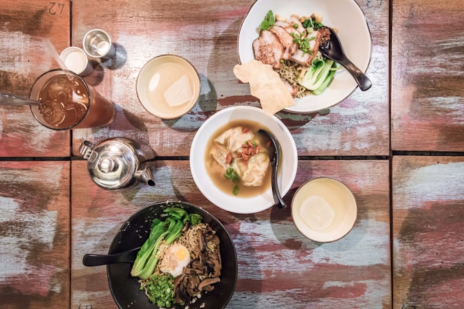An overhead view of a table with various dishes, including noodles with roasted pork, bok choy, and an egg. Nearby, there is a bowl of soup with dumplings. A glass of iced tea, a teapot, and several small bowls and cups are also arranged on the wooden surface.