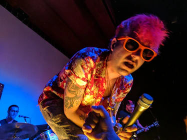 A performer with bright pink hair and orange sunglasses leans forward while holding a microphone. He is dressed in a colorful, floral-patterned shirt and has visible tattoos on his arms. In the background, a drummer plays a drum set, and another musician is holding a guitar.