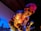 A performer with bright pink hair and orange sunglasses leans forward while holding a microphone. He is dressed in a colorful, floral-patterned shirt and has visible tattoos on his arms. In the background, a drummer plays a drum set, and another musician is holding a guitar.
