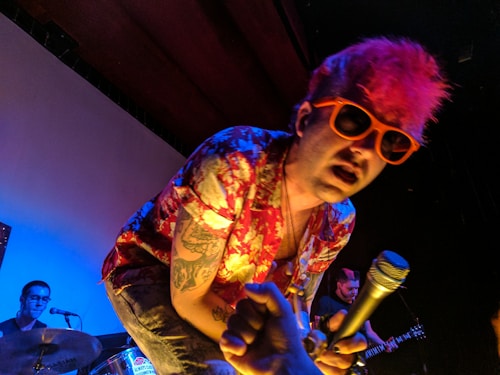 A performer with bright pink hair and orange sunglasses leans forward while holding a microphone. He is dressed in a colorful, floral-patterned shirt and has visible tattoos on his arms. In the background, a drummer plays a drum set, and another musician is holding a guitar.
