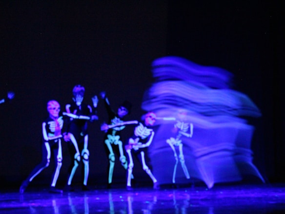 Several figures are dressed in black costumes with glowing skeleton designs, illuminated under ultraviolet light. The scene has a dynamic, blurred motion on the right side, suggesting movement or dance.