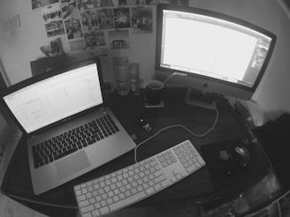 A workspace on a wooden desk with a laptop and a desktop monitor, both displaying white screens. A white keyboard and a computer mouse rest on the desk, along with a mug and several empty glasses. Numerous photographs and notes are attached to the wall behind the desk, adding a personal touch to the workspace.