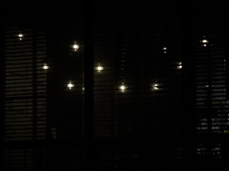Several dim lights are visible through a set of horizontal blinds, creating a pattern of illuminated spots against a shadowy background.