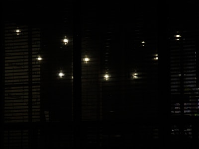 Several dim lights are visible through a set of horizontal blinds, creating a pattern of illuminated spots against a shadowy background.