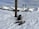 A snowy mountain landscape with snowshoes and trekking poles placed in the foreground near a signpost. The mountain range in the background is covered with snow under a partly cloudy sky.