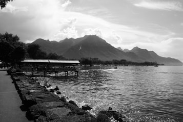 A serene lakeside scene featuring a promenade with a stone walkway, adjacent to a calm body of water. On the left, trees provide shade, and a wooden structure labeled 'H&ocirc;tel Restaurant' extends over the water. In the background, majestic mountains rise under a partially cloudy sky, creating a picturesque landscape.
