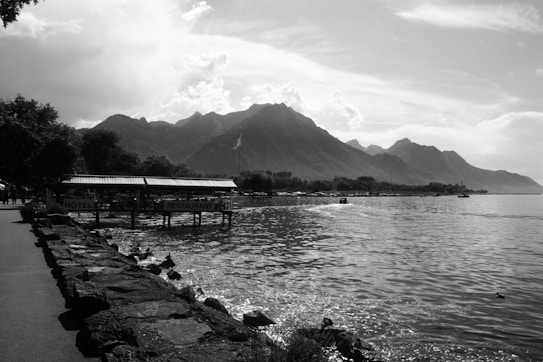 A serene lakeside scene featuring a promenade with a stone walkway, adjacent to a calm body of water. On the left, trees provide shade, and a wooden structure labeled 'H&ocirc;tel Restaurant' extends over the water. In the background, majestic mountains rise under a partially cloudy sky, creating a picturesque landscape.