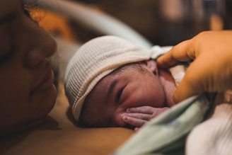 A newborn baby wearing a white knit cap lies on a woman's chest, with a hand gently touching the baby's head. The scene conveys intimacy and warmth.