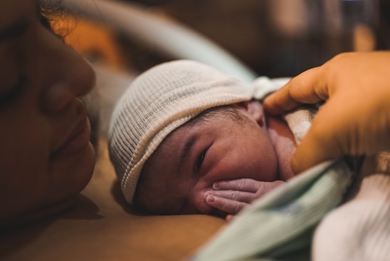 A newborn baby wearing a white knit cap lies on a woman's chest, with a hand gently touching the baby's head. The scene conveys intimacy and warmth.