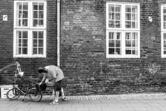 A person wearing a striped shirt and cap is standing next to a bicycle, adjusting or examining it. The scene is set against a brick wall with large windows. The overall atmosphere is calm and reflective, typical of an urban environment.
