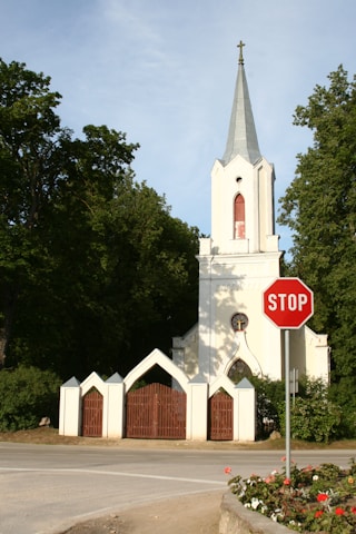 A white church with a tall steeple and a cross at the top stands amidst lush green trees. In the foreground, there is a red stop sign on a metal post by the road. The church features a gated entrance with red wooden doors, and there are flowers planted near the street.