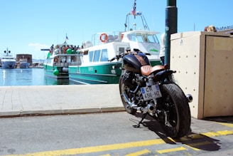 A motorcycle is parked near a waterfront on a sunny day. In the background, a ferry is docked at the harbor, with several people visible on board. The sky is clear and blue, and the scene conveys a sense of leisure and travel.