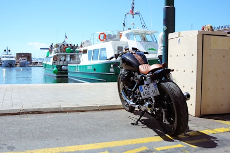 A motorcycle is parked near a waterfront on a sunny day. In the background, a ferry is docked at the harbor, with several people visible on board. The sky is clear and blue, and the scene conveys a sense of leisure and travel.