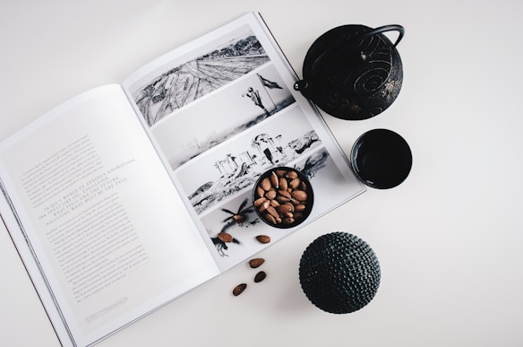 An open book showing black and white photographs and text is placed on a white surface. Surrounding the book are a black teapot, a small black cup, a bowl of almonds, and a textured black spherical object.