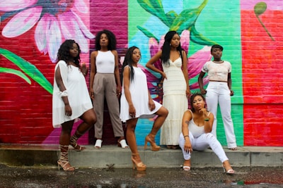 Six women pose confidently in front of a vibrant, colorful mural depicting large floral designs. The background consists of pink, green, and red hues. Each woman is dressed in stylish, contemporary outfits, ranging from white dresses to casual tops and pants. Their expressions and poses convey a sense of strength and empowerment.