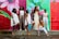 Six women pose confidently in front of a vibrant, colorful mural depicting large floral designs. The background consists of pink, green, and red hues. Each woman is dressed in stylish, contemporary outfits, ranging from white dresses to casual tops and pants. Their expressions and poses convey a sense of strength and empowerment.