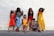 A group of six women pose against a light-colored brick wall. They are dressed in a variety of colorful and fashionable outfits. The women are positioned in a relaxed and confident manner, showcasing different styles and expressions.