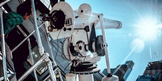 A large white telescope is mounted on a platform, pointing towards the sky on a sunny day. A child in a green striped shirt is leaning against a railing, observing the telescope. The sunlight creates a lens flare, and several buildings can be seen in the background.