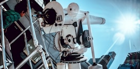 A large white telescope is mounted on a platform, pointing towards the sky on a sunny day. A child in a green striped shirt is leaning against a railing, observing the telescope. The sunlight creates a lens flare, and several buildings can be seen in the background.